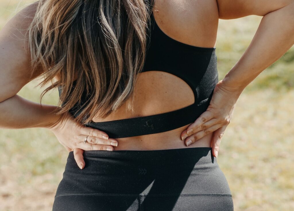 Spor kıyafetiyle beline elini koymuş sırt ağrısı çeken kadın, büyük göğsün omurgaya yükü